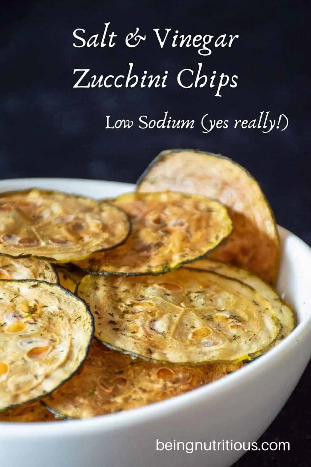 Zucchini chips in a white bowl in front of dark background.
