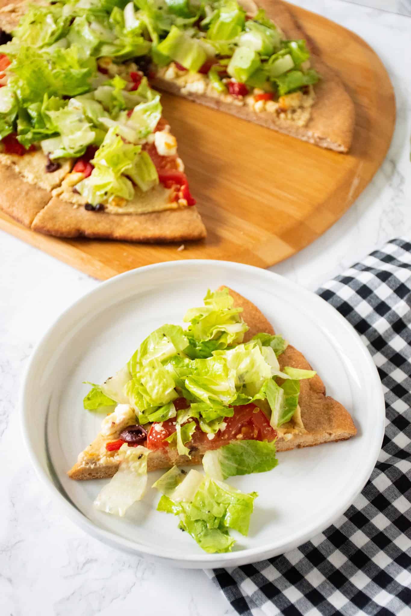 Slice of pizza on a plate. In background is the rest of the pizza on a peel, with whole wheat crust, topped with salad.