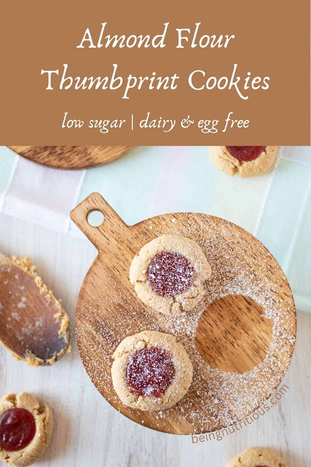 Two cookies on a wooden platter, sprinkled with powdered sugar. Text overlay: Almond flour thumbprint cookies; low sugar, dairy and egg free.