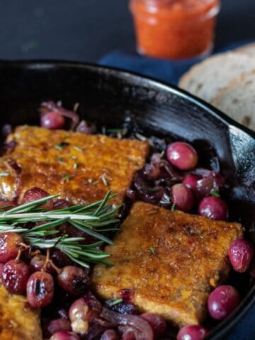 Cast iron skillet with tempeh, caramelized onions, and grapes in it. Garnished with fresh rosemary.