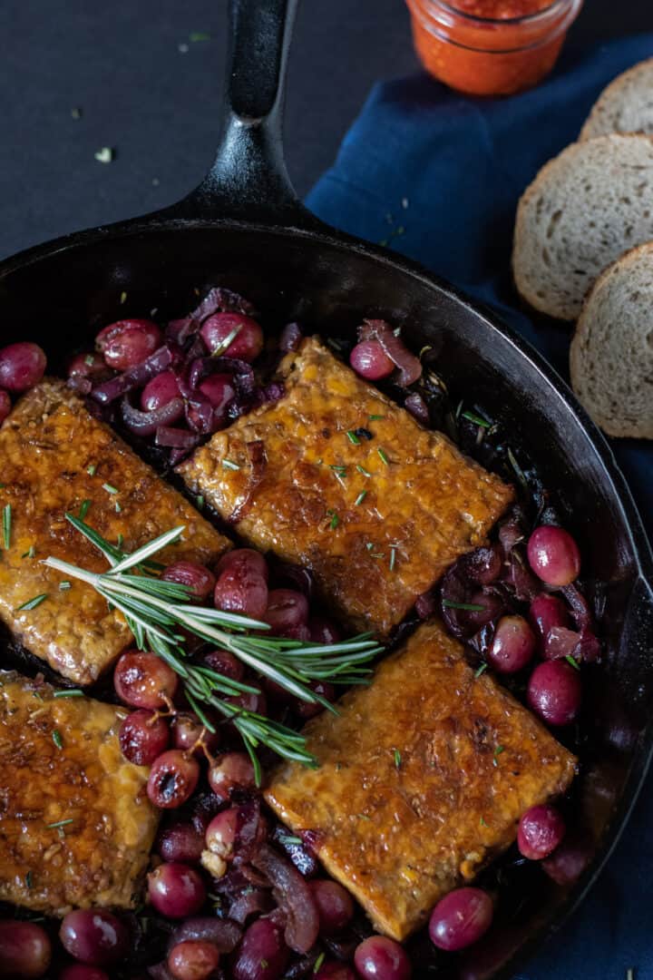 Cast iron skillet with tempeh, caramelized onions, and grapes in it. Garnished with fresh rosemary.