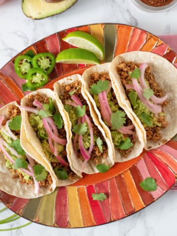 Five tacos lined up on a plate, surrounded by fixings.