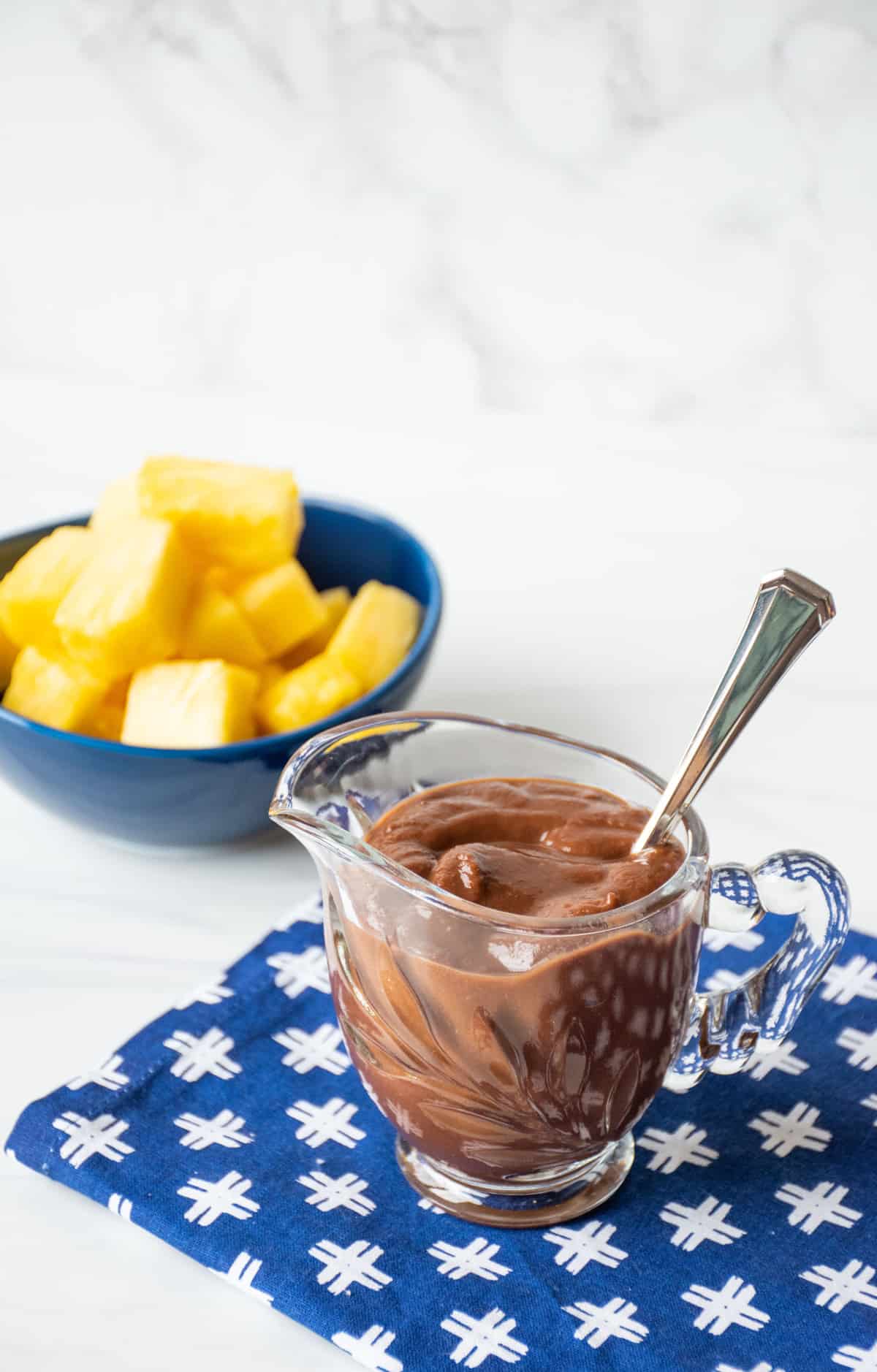 Chocolate sauce in a small glass pitcher, with a bowl of chopped pineapple in the background.