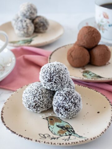 Homemade cherry truffles covered with shredded coconut, stacked on a small dish. Additional dishes with truffles covered with cocoa powder and ground almonds in the background, as well as 2 cups of coffee.