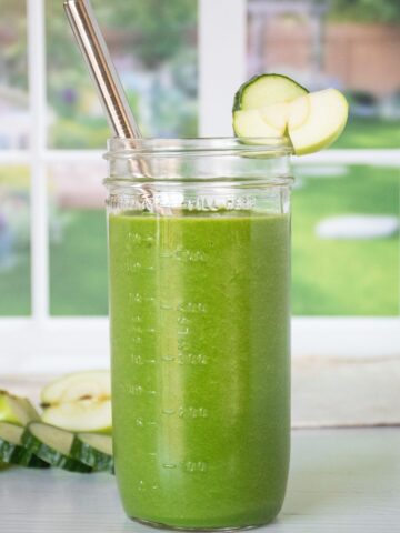A green smoothie in a tall Mason jar with a metal straw. Cucumber slices and green apple pieces in the background.