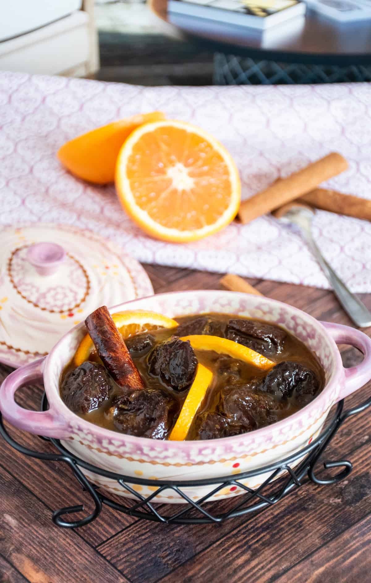Small crock with prune compote inside it, garnished with small slices of orange and a cinnamon stick.