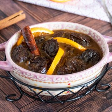 Small crock with prune compote inside it, garnished with small slices of orange and a cinnamon stick.