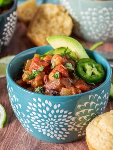 Bowl of chili, garnished with avocado, jalapeno slices, and cilantro, and a cornbread muffin on the side. Lime slices and additional bowls and spoons visible in the background.
