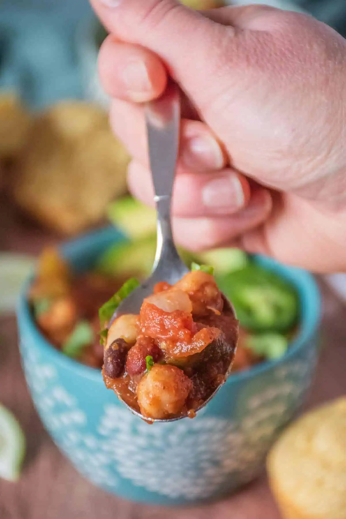 A hand holding a spoon with three bean chili on it. Bowl of chili visible in the background.