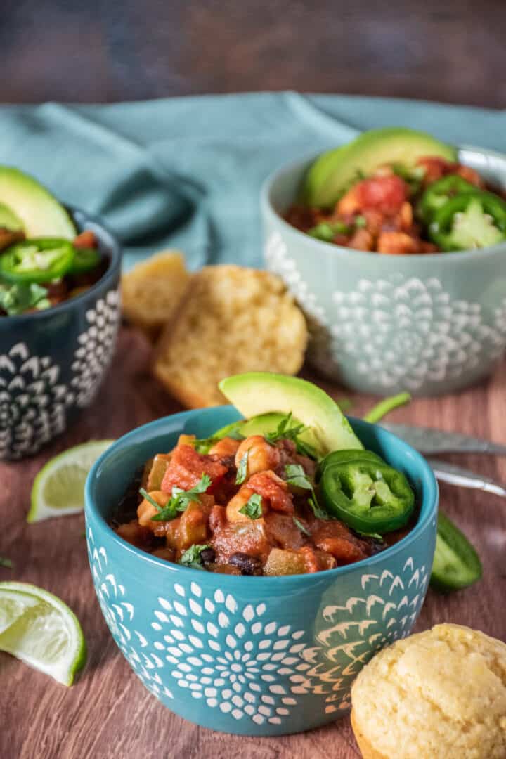 Bowl of three bean chili, garnished with avocado, jalapeno slices, and cilantro, and a cornbread muffin on the side. Lime slices and additional bowls and spoons visible in the background.