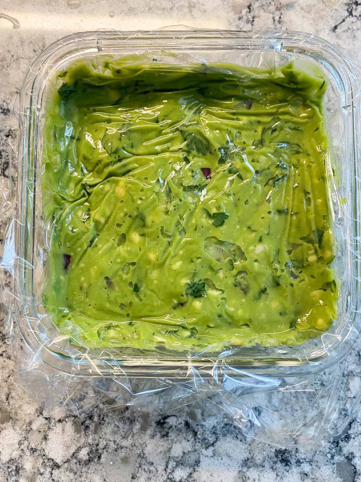 Guacamole in a glass container, with plastic wrap resting directly on top of the guacamole to prevent browning.