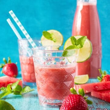 Glasses of agua fresca, with strawberries, watermelon slices, limes and mint scattered around. Carafe of beverage in background.
