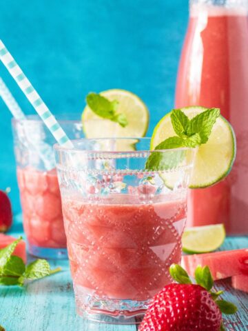 Glasses of agua fresca, with strawberries, watermelon slices, limes and mint scattered around. Carafe of beverage in background.