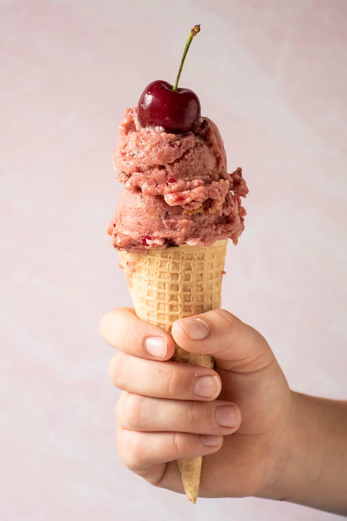 A hand holding an ice cream cone filled with cherry ice cream, and a cherry on the top.