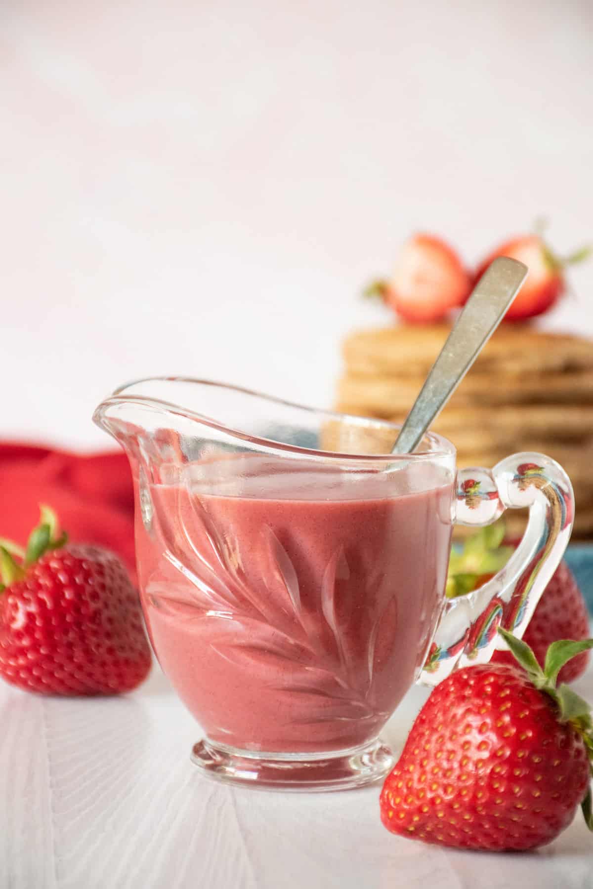 Strawberry sauce in a small glass creamer, with fresh strawberries scattered around.