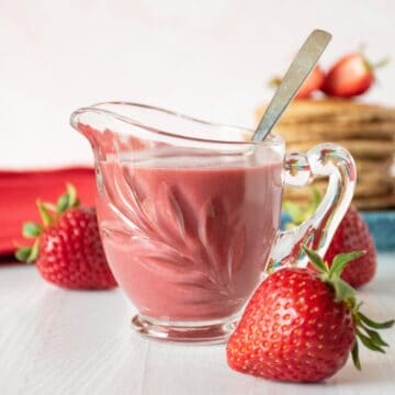 Strawberry sauce in a small glass creamer, with fresh strawberries scattered around.
