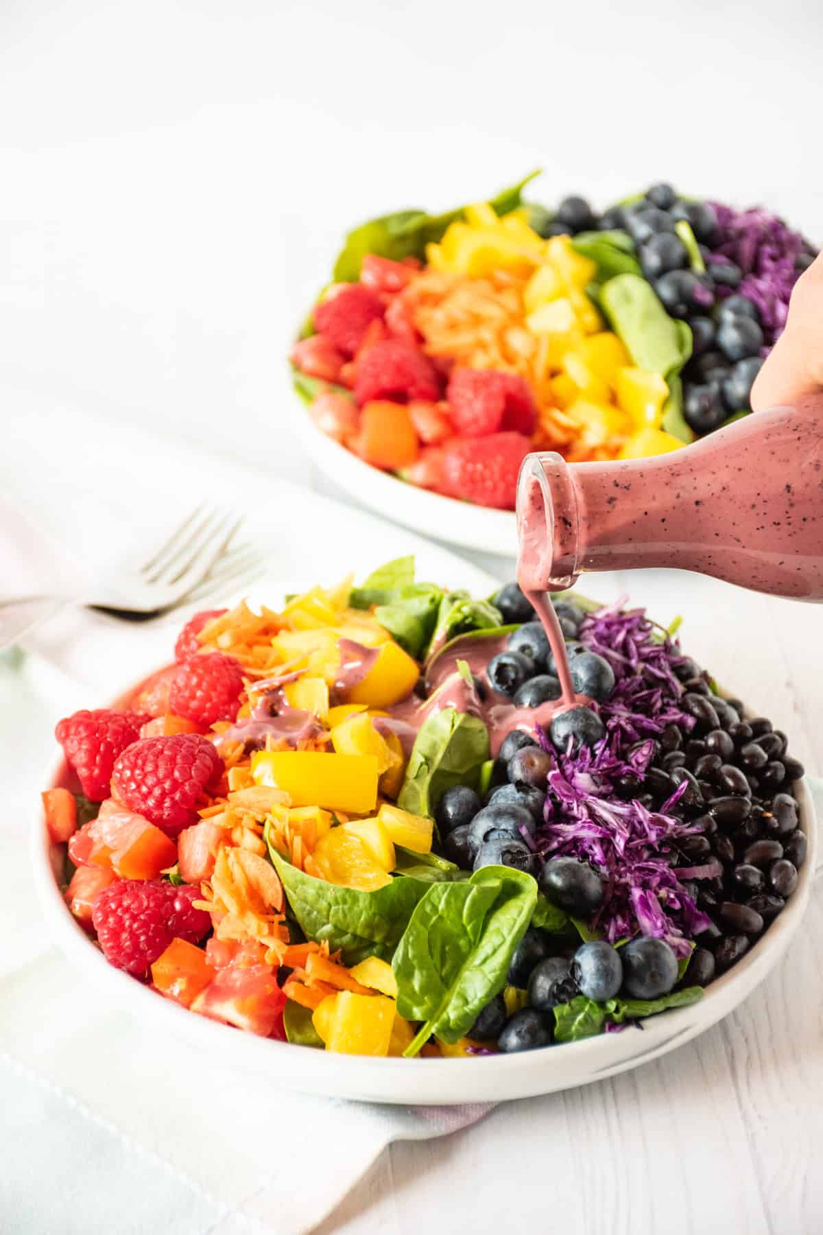 Salad with rainbow colored vegetables, with blueberry dressing being poured on it.