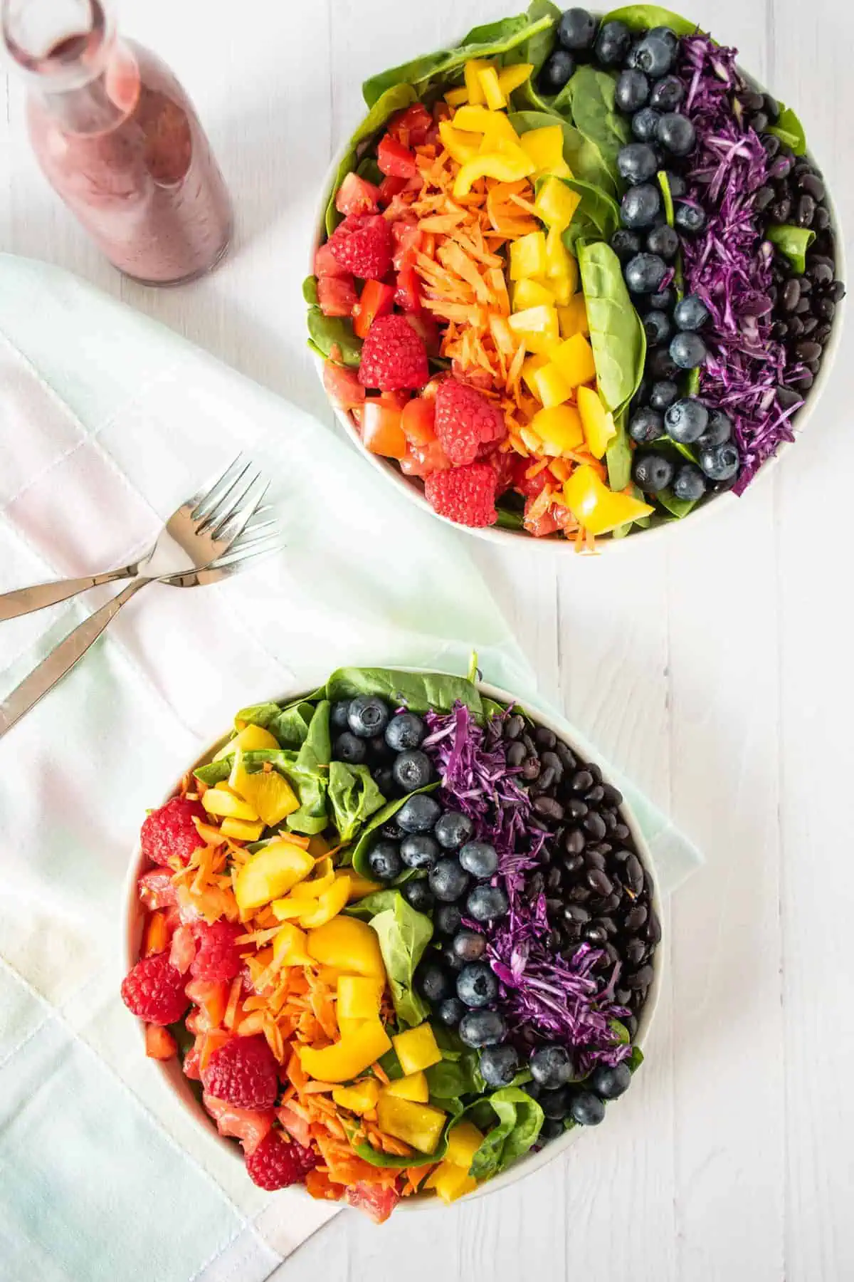 Two plates of salad with rainbow colored vegetables, and a bottle of blueberry dressing.
