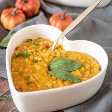 Pumpkin steel cut oats risotto in a heart shaped bowl, garnished with fresh sage leaves and chopped parsley.