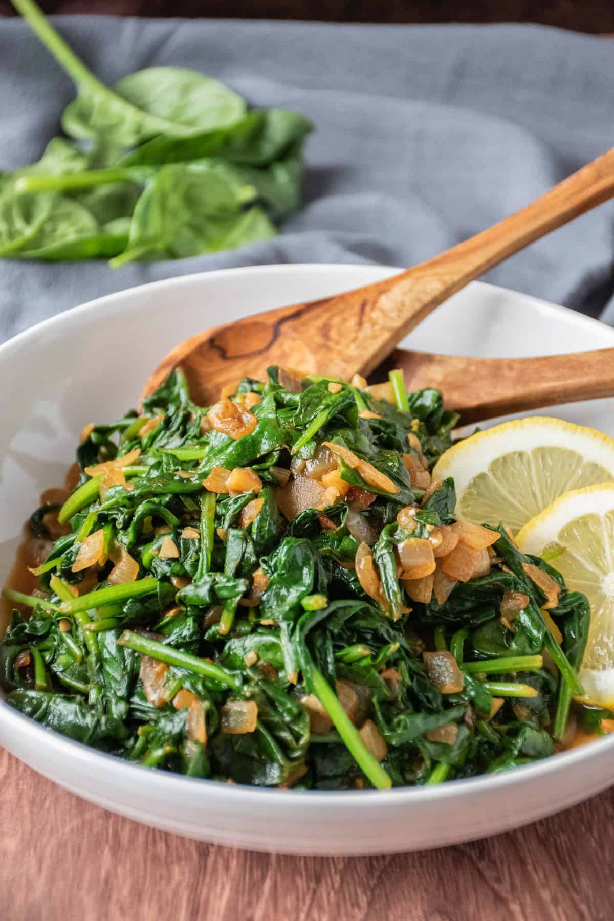 Serving bowl with sauteed spinach in it, with wooden serving spoons and garnished with lemon slices.