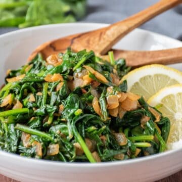 Serving bowl with sauteed spinach and onions in it, with wooden serving spoons and garnished with lemon slices.