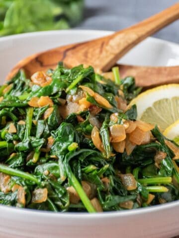 Serving bowl with sauteed spinach and onions in it, with wooden serving spoons and garnished with lemon slices.