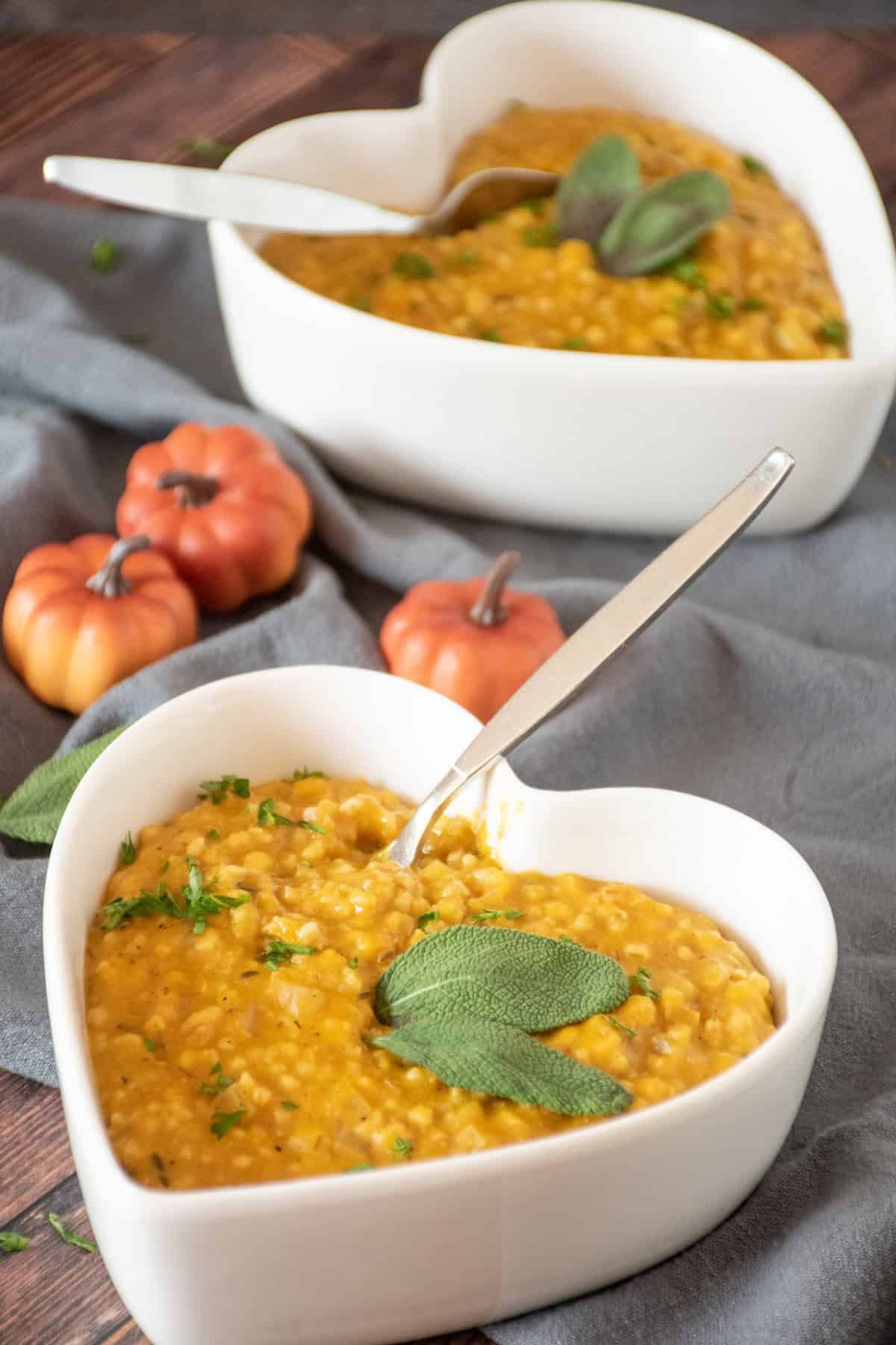 Pumpkin steel cut oats risotto in a heart shaped bowl, garnished with fresh sage leaves and chopped parsley.