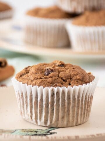 Single muffin on a dish. A stack of muffins is visible in the background.