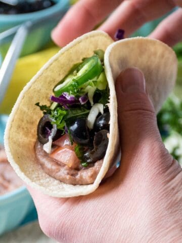 Close up image of 2 hands holding a taco, filled with refried beans, tomatoes, black olives, crunchy greens, and a jalapeno slice.