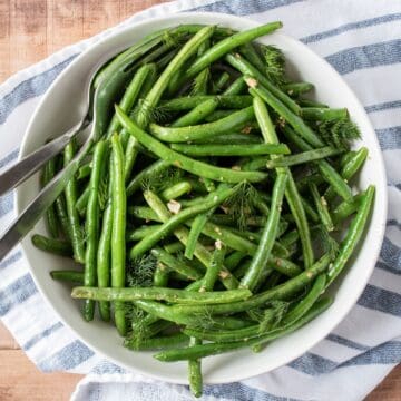 Roasted green beans in a bowl with serving utensils, garnished with fresh dill.