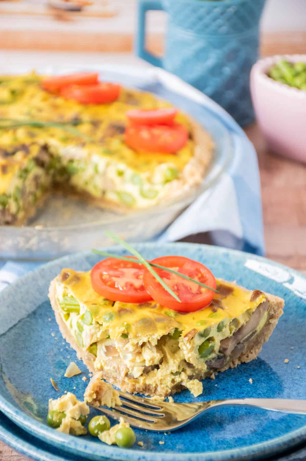 One slice of quiche on a plate, garnished with tomato slices and chives. A few bites taken from the front corner, and a fork lying on the dish.