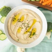 Bowl of creamy hummus, garnished with an olive oil drizzle, and a sprinkle of parsley, black sesame seeds, and sumac.