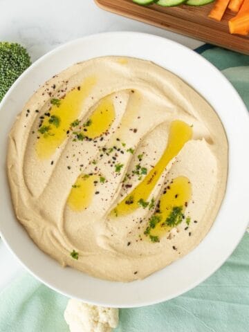 Bowl of creamy hummus, garnished with an olive oil drizzle, and a sprinkle of parsley, black sesame seeds, and sumac.