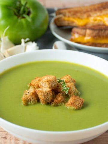 Green soup in a bowl, garnished with homemade croutons and fresh thyme. A grilled cheese sandwich and fresh garlic and green tomatoes are in the background.