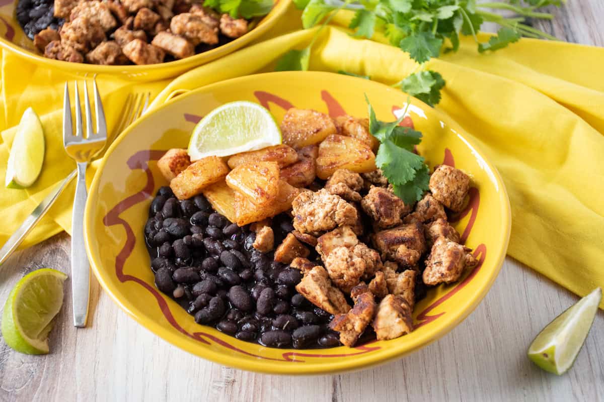 Bowl with jerk tofu and pineapple, over black beans, garnished with cilantro and a lime wedge.