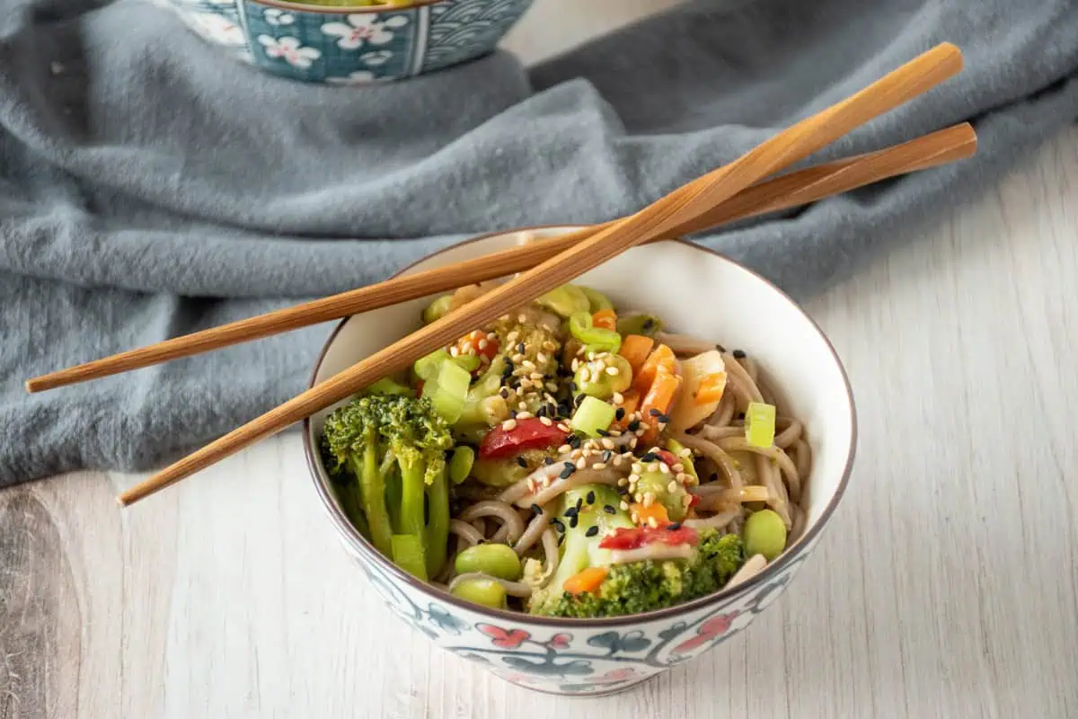 Bowl of noodles and vegetables, garnished with black and white sesame seeds and chopped green onions. A pair of chopsticks are laying across the bowl.