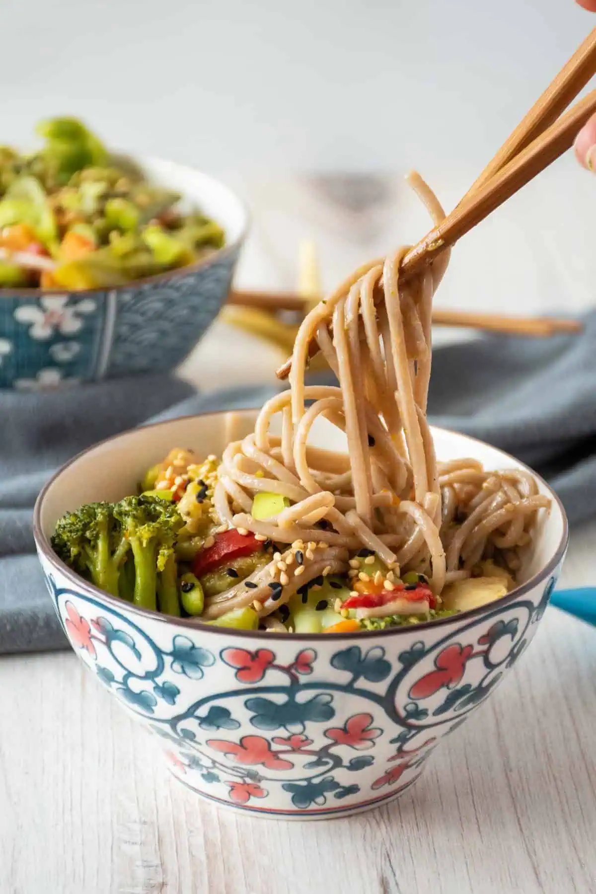 Bowl of noodles and vegetables, with some noodles being lifted from the bowl with chopsticks.