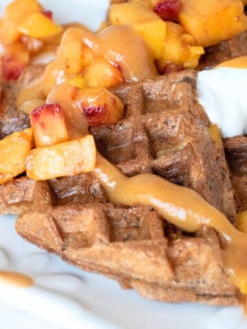 Waffle quarters, arranged on a serving dish, and garnished with chopped fresh peaches, dollops of yogurt, and peach syrup.