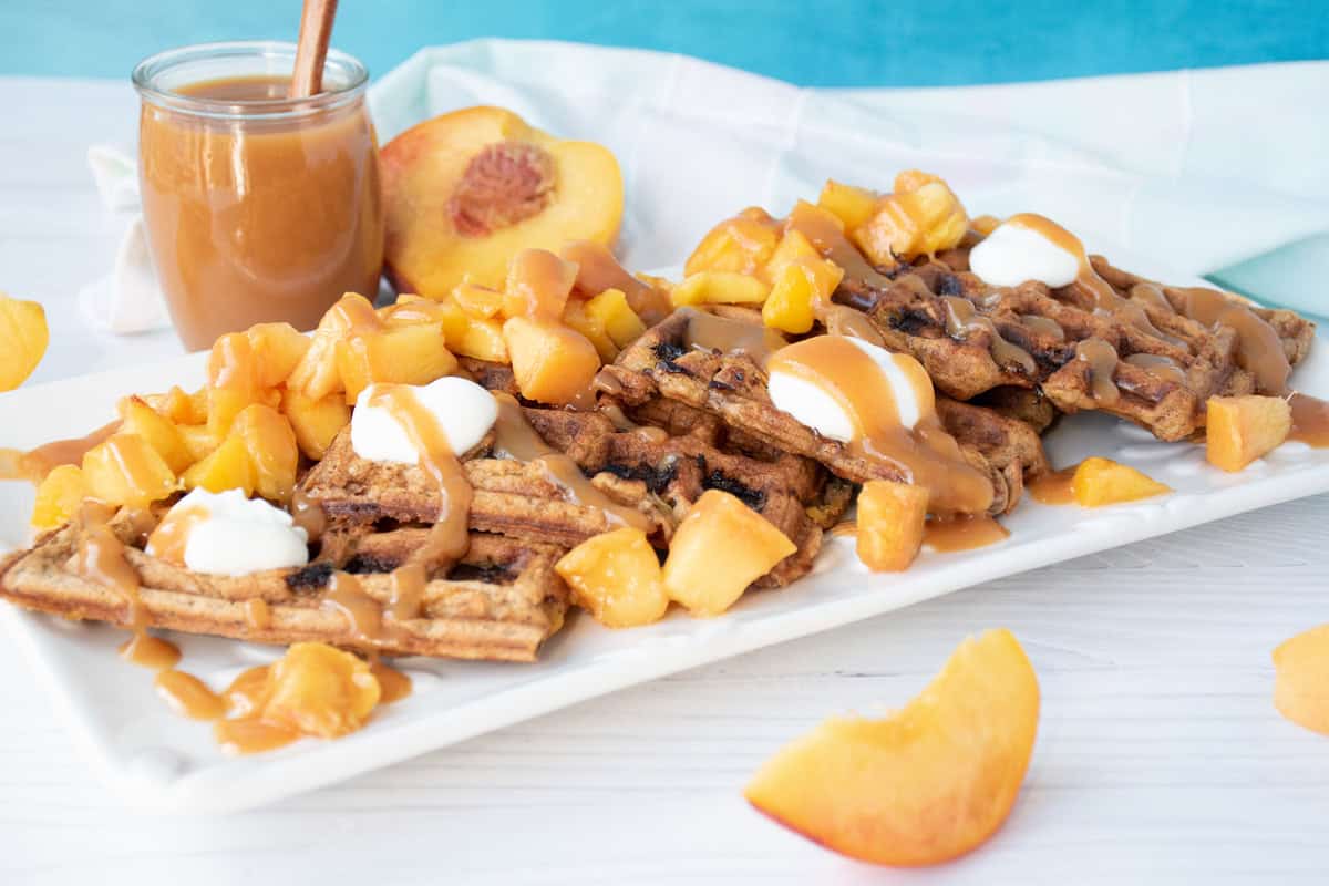 Waffle quarters, arranged on a serving dish, and garnished with chopped fresh peaches, dollops of yogurt, and peach syrup.