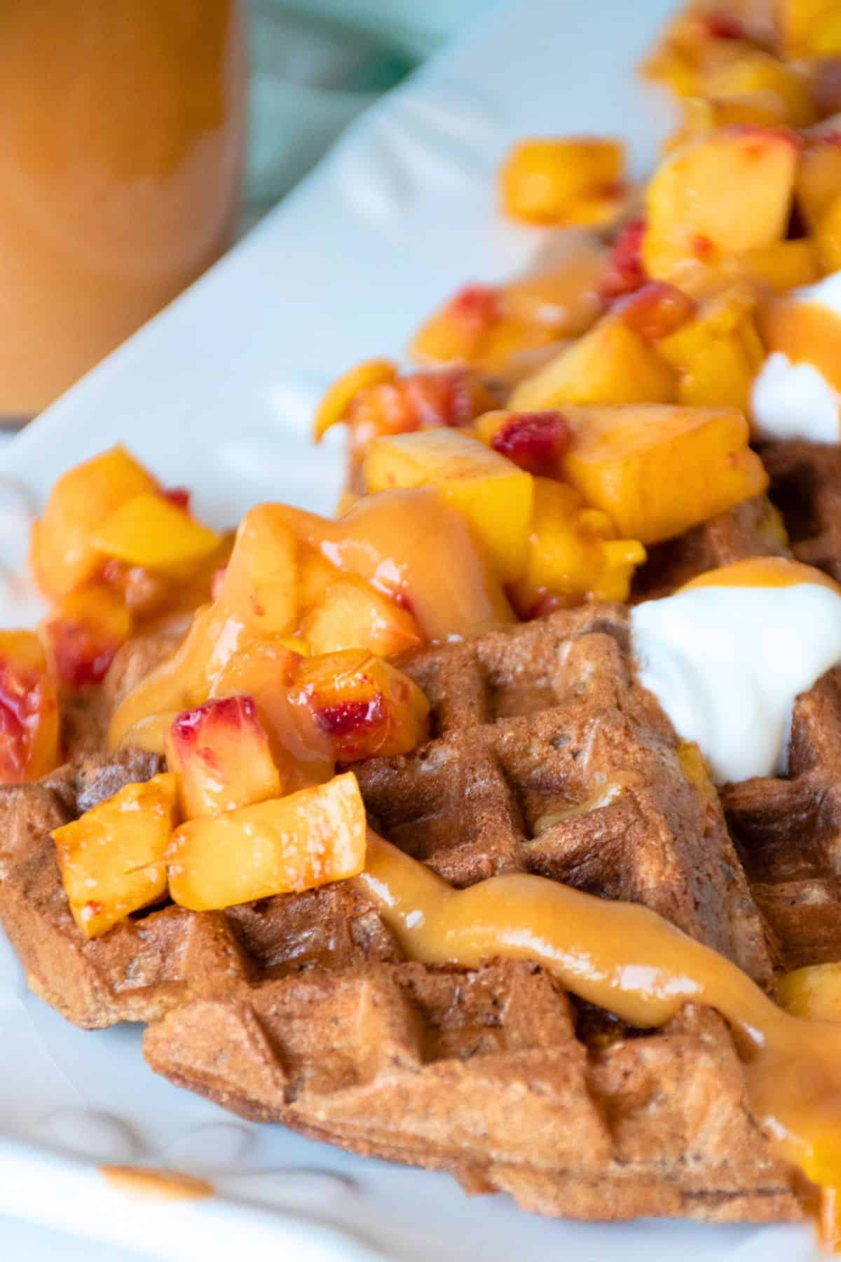 Waffle quarters, arranged on a serving dish, and garnished with chopped fresh peaches, dollops of yogurt, and peach syrup.