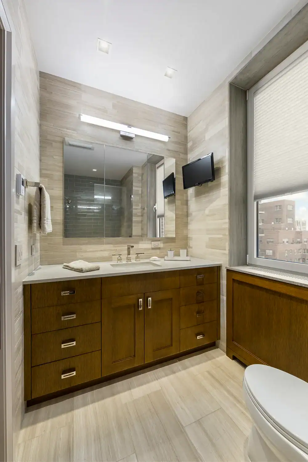 Contemporary bathroom with wooden vanity, large mirror, and window view.