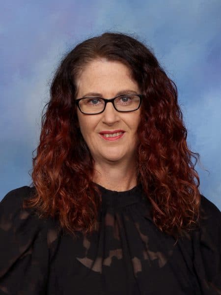 Professional woman with red curly hair and glasses, wearing a black top, against a blue background.