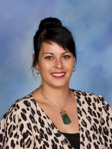 Bright young woman smiling, wearing animal print top, necklace, earrings, dark hair in bun, against blue background.