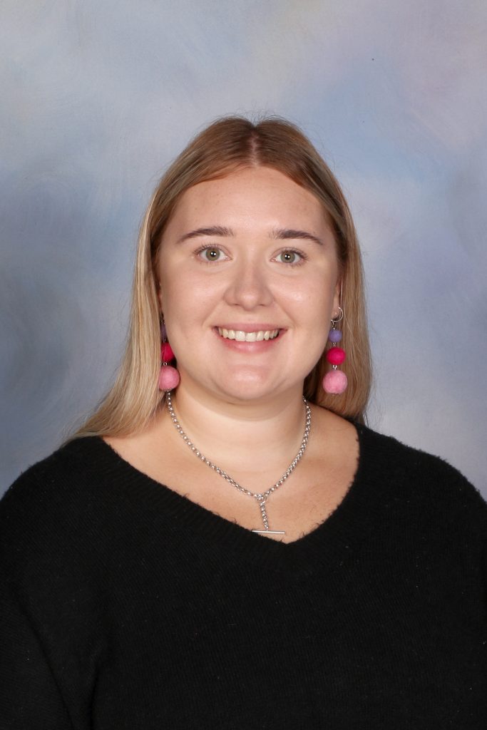 Bright smiling woman with long blonde hair and colorful pom-pom earrings, wearing a black top, portrait for Bell Block School.