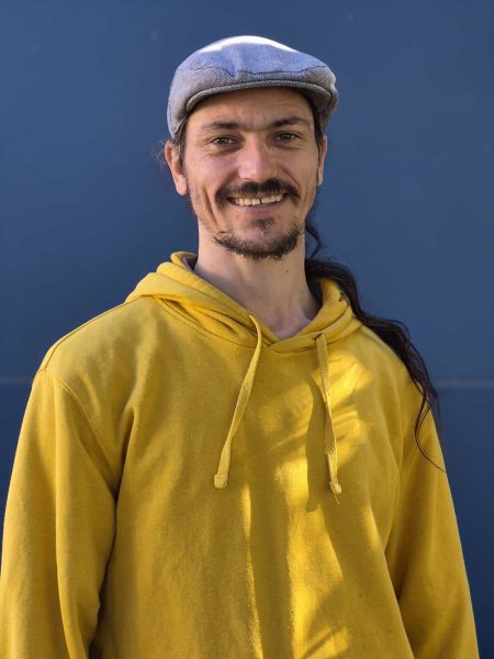 Staff member smiling in a yellow hoodie and grey cap at Bell Block School, New Zealand.