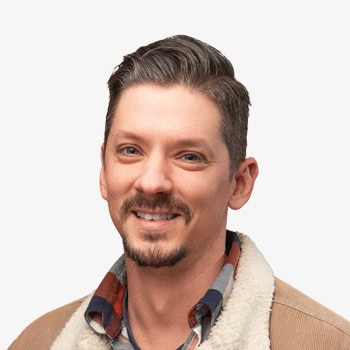 Smiling man with dark hair and beard, casual jacket, in natural setting.