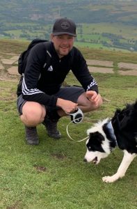 Robbie Harrison crouching with a Border Collie on a grassy hillside, holding a leash, with scenic co.