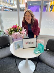 A woman smiling at a table with flowers, framed certificate, and office supplies in a modern office.