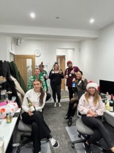 Group of colleagues celebrating Christmas in an office, wearing festive hats and holding drinks.