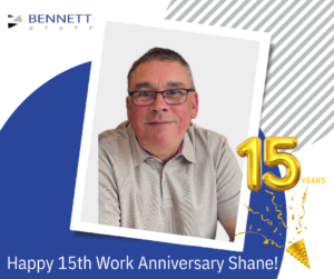 Portrait of Shane, smiling, wearing glasses and a light grey shirt.